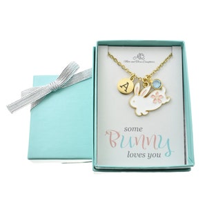 May include: A light blue gift box with a white bunny charm necklace inside. The necklace has a gold chain, a small round charm with the letter A, a light blue crystal, and a pink flower charm. The box has a white card inside with the text "some bunny loves you".