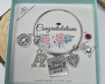 May include: A silver-toned expandable bangle bracelet featuring multiple charms including a compass, the letter A with a pink stone, a square charm engraved with Jesus loves me, a heart with a cross, a graduation cap, and a red stone.