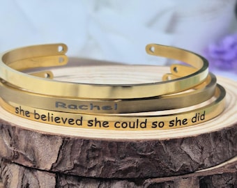 May include: Three gold-colored metal cuff bracelets stacked on a wooden slice. One bracelet is engraved with the name "Rachel", and another reads "she believed she could so she did" in black lettering. The bracelets are displayed against a soft background.