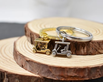 May include: Two golf cart keychains, one gold-toned and one silver-toned, rest on a wooden slice. The keychains feature detailed golf cart designs with wheels and interior elements. The background is a blurred wooden surface.