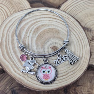 May include: Silver-toned graduation charm bracelet with multiple dangles. Charms include a pink owl, a graduation cap, a tassel, a pink gem, and the number "2026". The bracelet is displayed on a wooden surface.