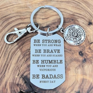 May include: A silver-colored keychain with a rectangular tag engraved with motivational phrases: "BE STRONG," "BE BRAVE," "BE HUMBLE," and "BE BADASS." It includes a U.S. Coast Guard emblem charm and a clasp.