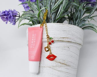 May include: A pink lip balm tube with the text "SUMMER FRIDAYS" and a gold chain charm. The charm features red cherries, a gold heart, and a red lip pendant. The lip balm is 0.23 oz.