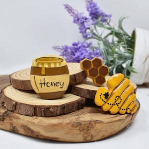 May include: Hand-painted wooden honey-themed decorations. Includes a barrel with "Honey" text, a honeycomb, and a bee. The items are yellow, brown, and black, set on wooden slices. Decorative items for a kitchen or home.