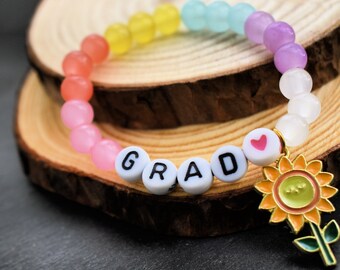 May include: A beaded bracelet with a rainbow gradient of colors. The bracelet has white letter beads that spell "GRAD" and a charm of a sunflower. The bracelet is on a wooden surface.