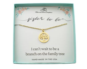 May include: A gold-colored necklace with a tree pendant, presented in a light blue box. The card inside reads "sister to be" and "I can't wait to be a branch on the family tree." The box is decorated with floral accents.