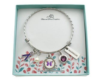 Könnte beinhalten: Ein silbernes Armband mit einem lila Band, einer Perle, einem Schmetterlingsanhänger, einem violetten Edelstein und einem Anhänger mit der Aufschrift "Warrior". Das Armband befindet sich in einer hellblauen Geschenkbox mit dem Aufdruck "Mom and Three Daughters" im Inneren.