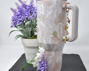 May include: A large, insulated tumbler with a gray lid and handle, adorned with a leaf pattern in muted tones. A gold charm with various pendants hangs from the handle. A potted lavender plant and sprigs of lavender and white flowers are in the background.