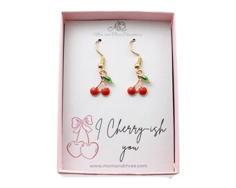 May include: A pair of gold-colored cherry earrings with red cherries and green leaves, presented in a pink box. The box features the text "I Cherry-ish you" and the website address www.momandthree.com.