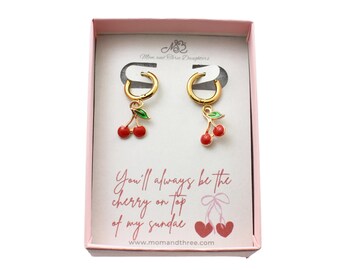 May include: Gold-tone hoop earrings with red cherry charms and green leaves, presented in a pink box. The box features the text "You'll always be the cherry on top of my sundae" and the website address.