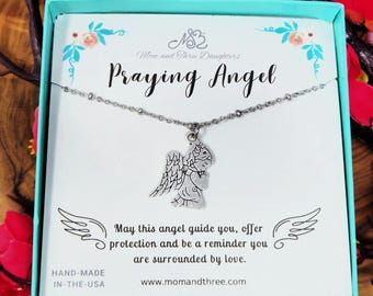 May include: A silver-colored necklace with a praying angel pendant, presented in a light blue gift box. The card inside reads "Praying Angel" and includes the text "May this angel guide you..." and "HAND-MADE IN-THE-USA".