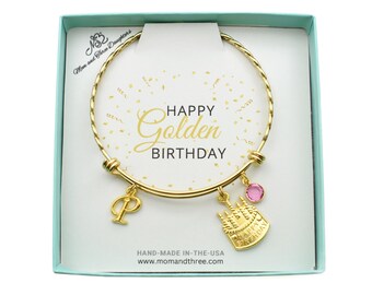 May include: A gold-colored bracelet with a charm that says "Happy Golden Birthday" in a gift box. The bracelet has a letter "P" charm, a pink gem, and a cake charm. The box is light blue with the text "Mom and Three Daughters".