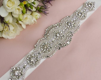 May include: A white satin bridal sash with a beaded embellishment featuring a crystal and pearl design.