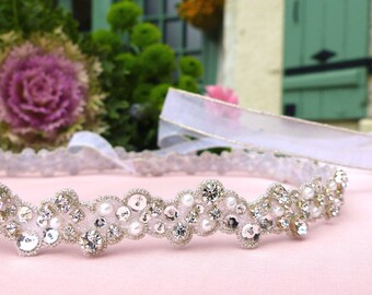May include: A delicate silver headband with a row of small white pearls and rhinestones. The headband is on a pink fabric background.