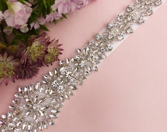Op de afbeelding: Een wit satijnen lint versierd met een rij glinsterende, heldere strass steentjes. De strass steentjes zijn in een bloemmotief gerangschikt.