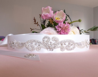 Op de afbeelding: Een witte satijnen bruidsband met een ontwerp van parels en strass. De band is versierd met een delicaat bloemenpatroon en glinsterende strass.