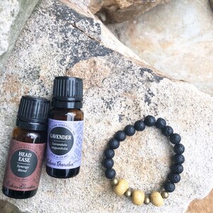 May include: Two small brown glass bottles with black caps and labels that read "Head Ease Synergy Blend" and "Lavender Lavandula angustifolia" with the brand name "Edens Garden". A black lava rock bracelet with gold beads is on a light brown rock surface.