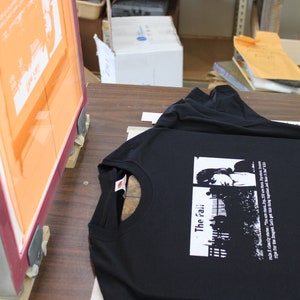 以下が含まれることがあります： 窓の前に立つ人の白のグラフィックプリントが施された黒いTシャツ。画像の下に「The 'rai'」というテキストが印刷されています。