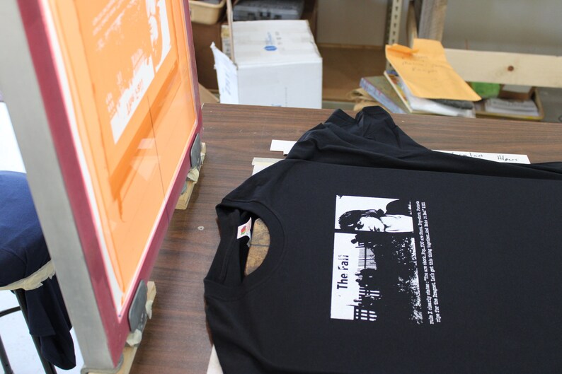 以下が含まれることがあります： 窓の前に立つ人の白のグラフィックプリントが施された黒いTシャツ。画像の下に「The 'rai'」というテキストが印刷されています。