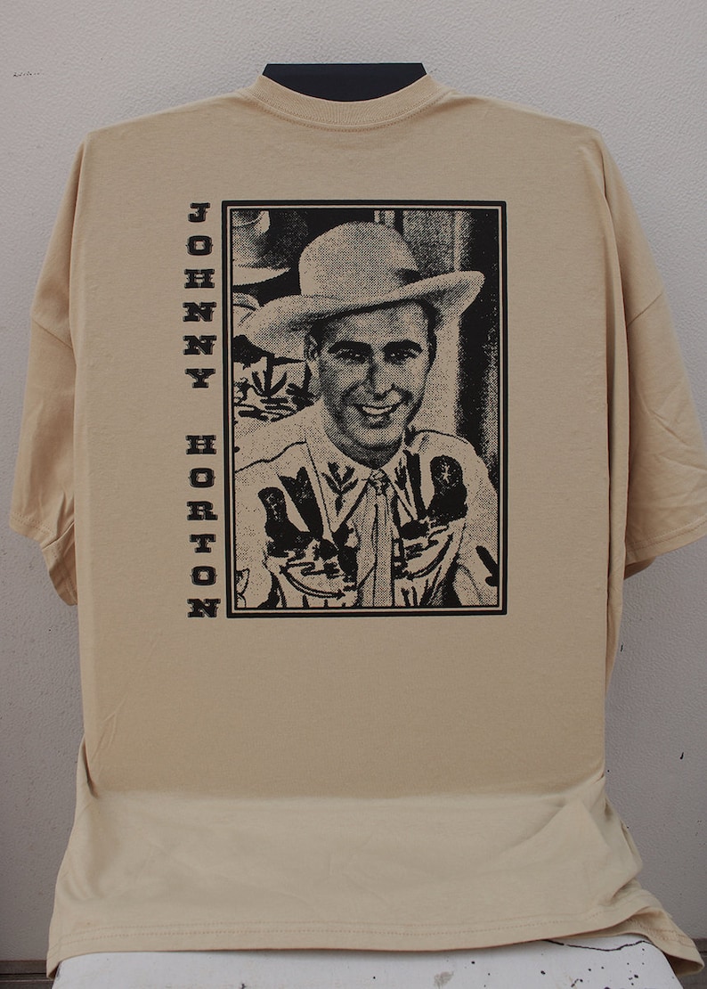 Może przedstawiać: Beżowa koszulka z czarno-białym portretem Johnny'ego Hortona w kapeluszu kowbojskim. Imię "JOHNNY HORTON" jest wydrukowane pionowo po lewej stronie czarnymi literami. Koszulka ma kr&oacute;tkie rękawy i okrągły dekolt.