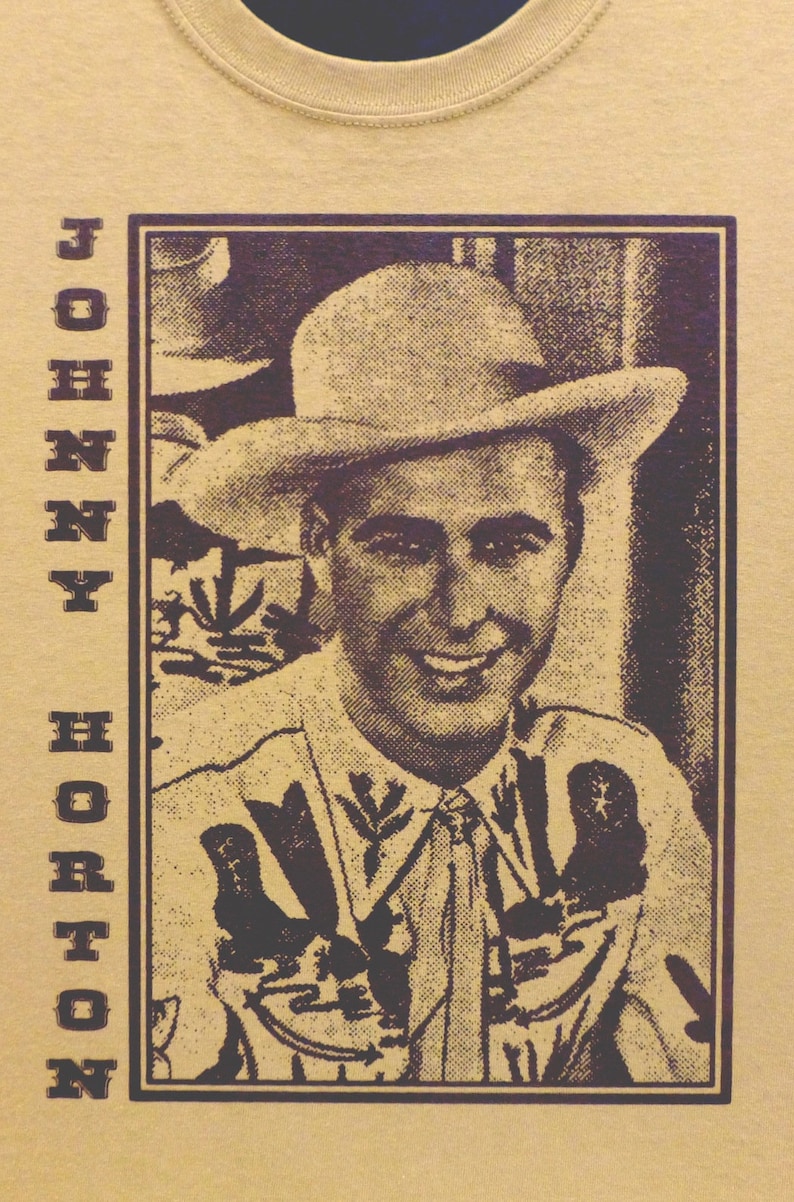 Puede incluir: Una camiseta de estilo vintage con una imagen en blanco y negro de Johnny Horton con un sombrero y una camisa estampada. El texto "JOHNNY HORTON" est&aacute; impreso verticalmente en el lado izquierdo de la imagen.