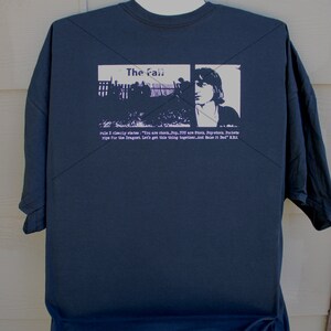 以下が含まれることがあります： 「ザ・フォール」というバンドの白いプリントが施された濃い青色のTシャツ。プリントには、人物と建物の白黒画像が含まれています。画像の下のテキストには、「rule X clearly states: 'You are chook. Pop. YOU are Stock. Pop-stock. Pockets ripe for the Dragnet. Let's get this thing together. And Make it Zed' KRA」と書いてあります。