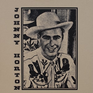Może przedstawiać: Beżowa koszulka z czarno-białym portretem mężczyzny w kapeluszu i koszuli kowbojskiej. Nazwisko "JOHNNY HORTON" jest wydrukowane pionowo po lewej stronie czarnymi literami.