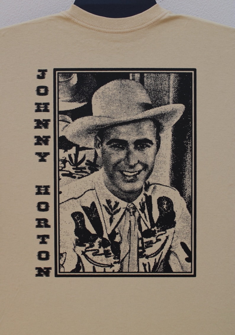 Może przedstawiać: Beżowa koszulka z czarno-białym portretem mężczyzny w kapeluszu i koszuli kowbojskiej. Nazwisko "JOHNNY HORTON" jest wydrukowane pionowo po lewej stronie czarnymi literami.
