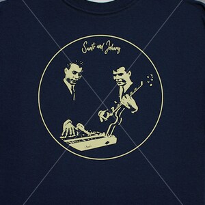 May include: A navy blue t-shirt with a circular design featuring a black and white illustration of two musicians playing instruments. The text "Santo and Johnny" is written in a cursive font above the musicians.
