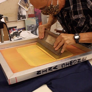 May include: A person uses a squeegee to spread yellow ink across a screen printing frame. The frame is resting on a blue t-shirt. The text "SHA SHEON M" is written on the frame.