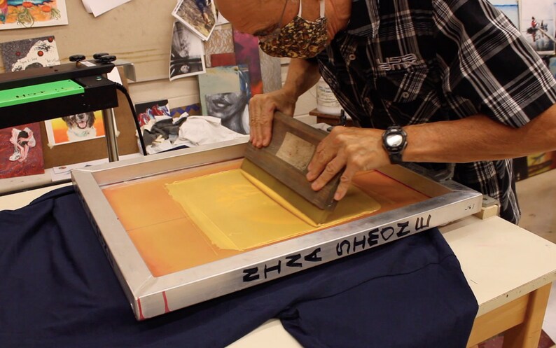 May include: A person uses a squeegee to spread yellow ink across a screen printing frame. The frame is resting on a blue t-shirt. The text "SHA SHEON M" is written on the frame.