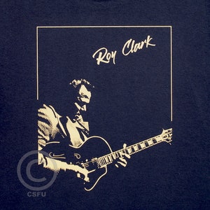 May include: A navy blue t-shirt with a white silhouette of a man playing a guitar. The text "Roy Clark" is written in white above the silhouette.