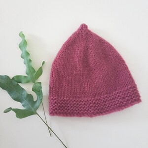 May include: A knitted hat in a deep pink colour. The hat has a simple design with a pointed crown and a folded brim.