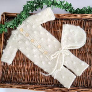 May include: A cream-colored knitted baby outfit set, including a long-sleeved button-up top, pants, and a bonnet. The set features a textured, dotted pattern and wooden buttons. The outfit is displayed on a woven wicker tray with greenery.