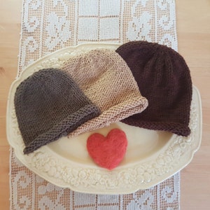 May include: Three knitted hats in brown, beige, and dark brown on a white plate with a red heart. The hats are all the same style, with a simple, rounded shape.