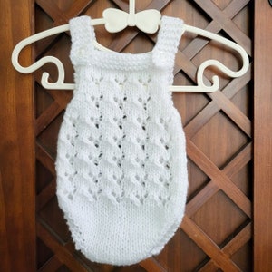 May include: A white knitted romper featuring a delicate openwork lace pattern throughout the body. The garment has two shoulder straps secured with small white buttons at the front and a ribbed neckline, all displayed on a white plastic hanger.