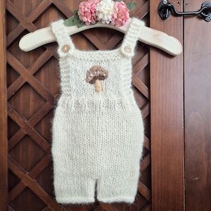 以下が含まれることがあります： 胸元に茶色のキノコの刺繍が施されたクリーム色のニットロンパース。小さな木製ボタンで留められた2本のショルダーストラップと、ふわふわとした質感の毛糸仕上げが特徴です。木製のハンガーに掛けられています。
