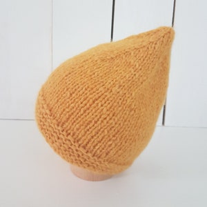May include: A mustard yellow knitted beanie hat with a pointed top. The hat is displayed on a wooden mannequin head.