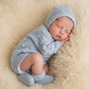 May include: A sleeping baby wearing a light blue knitted outfit, including a hat, romper, and booties. The outfit has a textured, dotted design. The baby is lying on a soft, cream-colored, fluffy surface.
