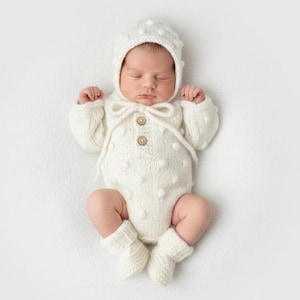 May include: A newborn baby wearing a white knitted outfit, including a bonnet, onesie, and booties. The outfit features a textured design with small, raised bobbles and wooden buttons. The baby is lying down with eyes closed.