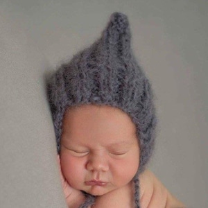 May include: A dark gray knitted bonnet with a pointed top, featuring a textured ribbed pattern and soft, fuzzy yarn construction. The headwear is designed with long ties that extend from the sides, providing a secure fit around the head.