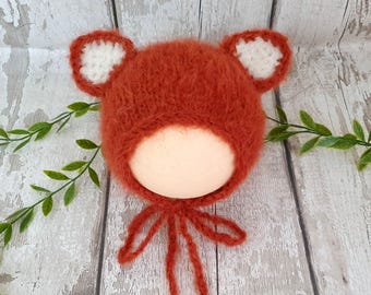 May include: A fuzzy, rust-orange knitted bonnet featuring two rounded ears with white centers. The bonnet has a textured, mohair-like appearance and includes two long, matching rust-orange yarn ties attached at the base for securing the item.