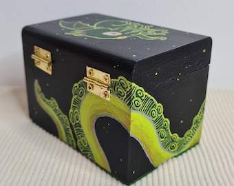 May include: A black wooden box with a green and yellow octopus design. The box has gold hinges and a black background with white stars.