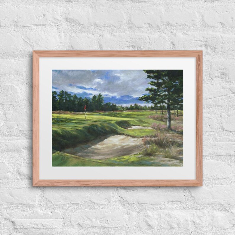 May include: A framed painting of a golf course with a green fairway, sand traps, and a red flag in the distance. The painting is set against a backdrop of a cloudy sky and trees.