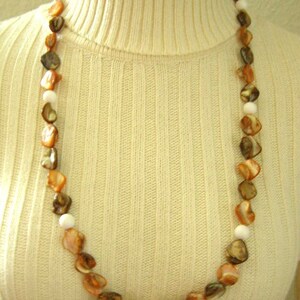 Può includere: Una lunga collana fatta di perline di conchiglia marroni e bianche. La collana è su un maglione a collo alto bianco.