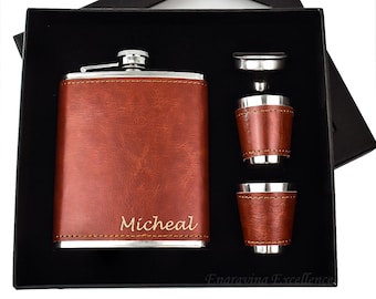 Puede incluir: Una petaca de acero inoxidable forrada de cuero marrón con un tapón plateado y dos vasos de chupito a juego. La petaca está grabada con el nombre "Micheal".