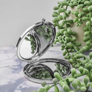 May include: A silver compact mirror with a hinged lid and two round mirrors. The mirror is open and the lid is propped up behind it.