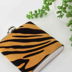 May include: A stainless steel hip flask with a tiger print velvet cover. The flask is lying on a white surface.