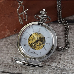 May include: A silver pocket watch with a white face, Roman numerals, and a visible gold-colored mechanical movement. The watch is open, revealing the inner workings. A silver chain and clasp are attached. The watch is labeled "CHARLES HENDON".