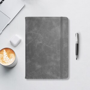 May include: A rectangular gray notebook with a textured cover and a vertical elastic closure band. The notebook is positioned centrally, accompanied by a silver ballpoint pen, a white wireless earbud case, and a cup of latte coffee.